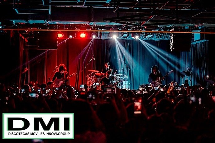Escenario de concierto con banda en directo y diseño de iluminación basado en potentes haces blancos verticales.