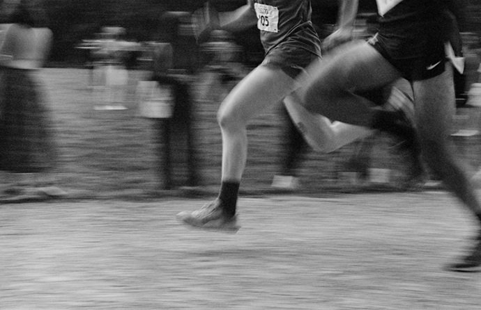 Imagen artística en blanco y negro con efecto de desenfoque de movimiento que muestra las piernas de varios atletas corriendo en una pista, representando el dinamismo y el esfuerzo deportivo.